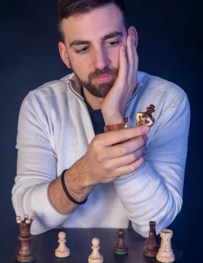Un homme en pull blanc contemple une pièce déchecs, la tête posée sur une main. Il est assis à une petite table sur laquelle sont posées diverses pièces déchecs. Larrière-plan est un dégradé sombre.