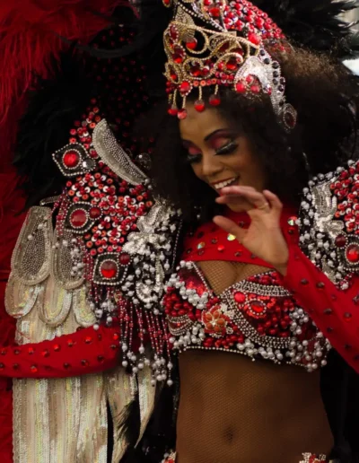 Une personne vêtue dun costume de carnaval coloré orné de plumes rouges et noires, de perles complexes et dune coiffe ornée de bijoux sourit avec délice. Le costume comporte des ailes élaborées et des ornements scintillants, capturant la joie de la célébration.