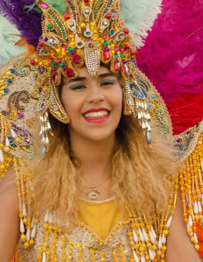 Une personne souriante, vêtue dun costume de carnaval vibrant et coloré orné de plumes et de perles, avec une coiffe élaborée aux accents dorés, rouges et violets. Larrière-plan est rempli de grandes plumes multicolores.