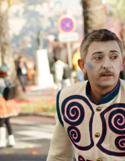 Une personne vêtue dun costume blanc aux motifs tourbillonnants colorés et au visage peint participe à un festival en plein air animé. Des silhouettes floues et des palmiers sont visibles en arrière-plan.