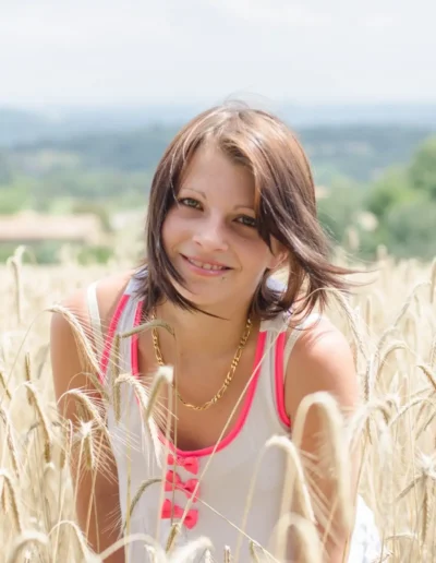 Une femme souriante aux cheveux bruns est accroupie dans un champ de blé doré par une journée ensoleillée. Elle porte un débardeur blanc avec des accents roses. Larrière-plan montre un paysage pittoresque avec un ciel brumeux.