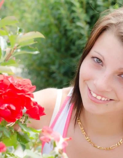 Une jeune femme aux cheveux bruns sourit à la caméra tout en tenant des roses rouges éclatantes dans un jardin. Elle porte un collier en or et un débardeur blanc. La verdure luxuriante lentoure, créant une atmosphère chaleureuse et sereine.