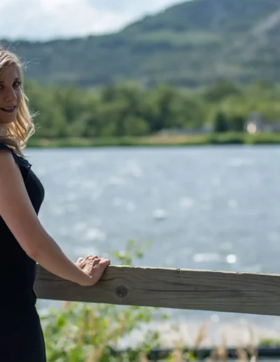 Une personne aux cheveux blonds et vêtue dune robe noire se tient debout près dune clôture en bois, surplombant un lac étincelant. Larrière-plan présente un paysage verdoyant et luxuriant avec des collines sous un ciel nuageux.