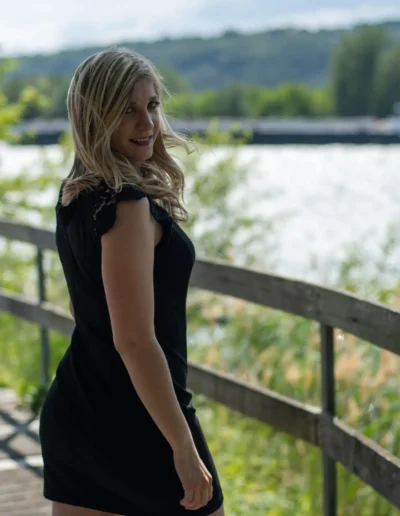 Une femme en robe noire se tient sur un chemin en bois près dun plan deau, entourée de verdure. Elle regarde par-dessus son épaule, avec des collines visibles en arrière-plan, sous un ciel partiellement nuageux.