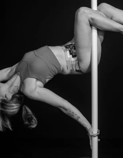 Dans cette photographie professionnelle en noir et blanc saisissante, une femme exécute un mouvement de pole dance, cambrant gracieusement son dos avec ses jambes enroulées autour de la barre. Vêtue dun soutien-gorge de sport et dun short, elle met en valeur à la fois son équilibre et sa force sur le fond sombre.