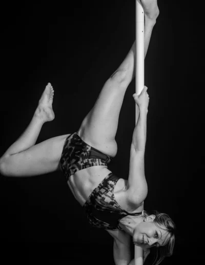 Une femme exécute un mouvement de pole dance, suspendue la tête en bas, tenant la barre avec un bras et une jambe enroulés autour. Elle est vêtue dune tenue de sport à motifs sur un fond sombre.