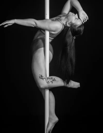 Une personne aux cheveux longs et portant un tatouage floral sur la jambe prend la pose sur un poteau sur un fond sombre. La pose comprend les bras et les jambes étendus, mettant en valeur la force et léquilibre.