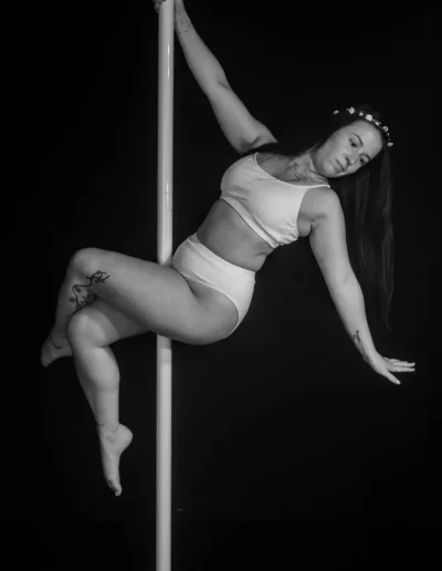 Une femme exécute une pole dance dans un studio, vêtue dune tenue de couleur claire et dun bandeau fleuri. Limage est en noir et blanc, mettant en valeur sa pose gracieuse et son expression. Des tatouages sont visibles sur son bras et sa jambe.