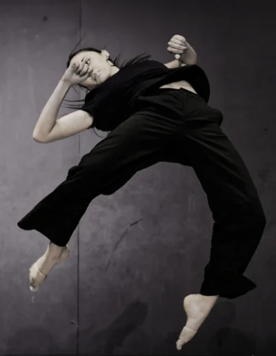 Une personne vêtue de noir exécute un saut de danse dynamique sur un fond sombre, les bras et les jambes étendus dans les airs, transmettant énergie et mouvement.