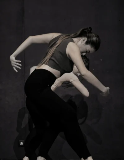 Deux danseurs exécutent des mouvements contemporains sur un fond sombre. La danseuse au premier plan, vêtue dun haut court et dun legging, est capturée dans une pose gracieuse et courbée. Les ombres ajoutent de la profondeur à la scène, soulignant la fluidité et lémotion de la danse.