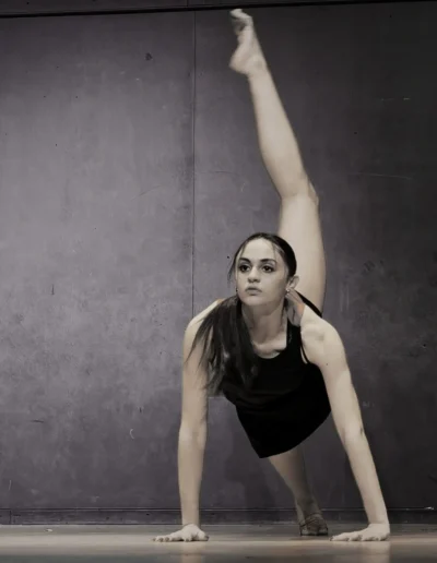 Une danseuse en justaucorps noir exécute une routine au sol, en équilibre sur les mains et un pied, lautre jambe tendue verticalement. Larrière-plan est un mur uni et texturé, créant une atmosphère austère et concentrée.