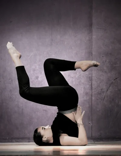 Une personne en tenue de sport est en équilibre sur ses épaules, les jambes pliées au niveau des genoux et les pieds levés, effectuant une pose de yoga sur un plancher en bois sur un fond sombre et texturé.