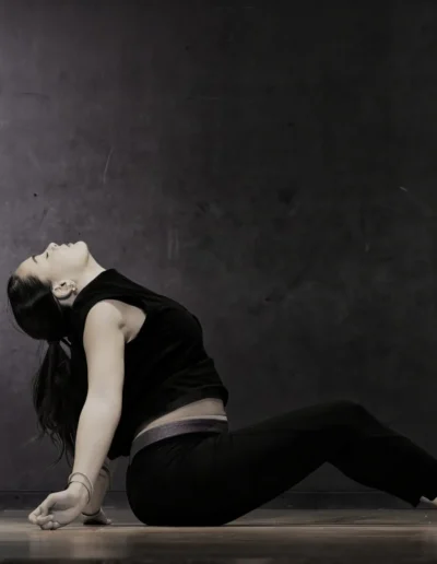 Une femme vêtue de vêtements sombres exécute une posture de yoga assise sur le sol, le dos cambré et la tête penchée en arrière, les mains posées derrière elle pour se soutenir. Larrière-plan est un mur uni et sombre.