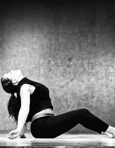 Une femme aux cheveux noirs, vêtue dun haut sans manches et dun legging, sappuie sur ses mains dans une posture de yoga assise. Elle cambre le dos et regarde vers le haut, appuyée contre un mur texturé dans une image en niveaux de gris.