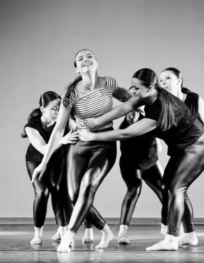Cinq danseuses se produisent sur scène dans une pose dramatique. Quatre danseuses en costumes noirs soutiennent ou entourent une danseuse centrale en haut rayé et jambières, qui semble exprimer une forte émotion. L'image est en noir et blanc.