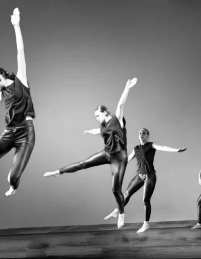 Quatre danseurs en tenue noire sautent et se déplacent énergiquement sur une scène, capturés en plein vol et dans des poses dynamiques sur un fond uni. L'image est en noir et blanc.