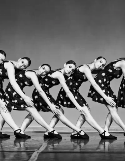 Six danseurs vêtus de robes à pois assorties et de chaussures noires prennent une pose synchronisée et penchée sur la scène, créant ainsi une ligne courbe. La photo est en noir et blanc.