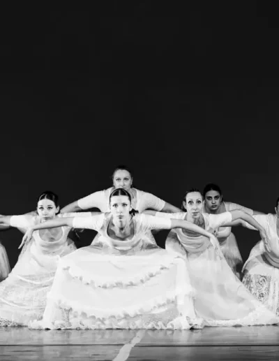 Sept danseuses vêtues de longues robes blanches fluides posent en ligne sur scène, les bras tendus et pliés aux coudes, créant une formation symétrique et dramatique sur un fond sombre.