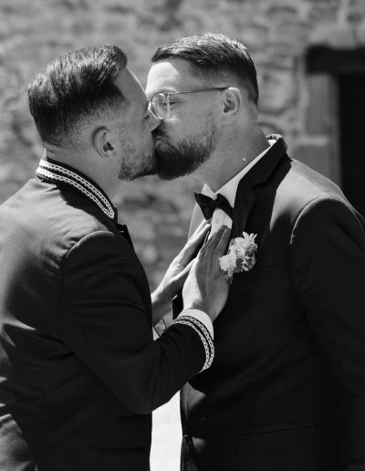 Deux hommes en costume s'embrassent en plein air devant un bâtiment en pierre, l'un avec une boutonnière et des lunettes. Le moment semble intime et joyeux, probablement dans le cadre d'un mariage ou d'une célébration spéciale.