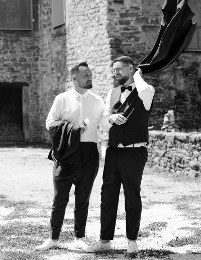 Deux hommes habillés de manière formelle se tiennent à l'extérieur, près d'un bâtiment en pierre. L'un d'eux ajuste sa veste sur son épaule, tandis que l'autre tient la sienne, et tous deux semblent en pleine conversation. La scène est en noir et blanc.