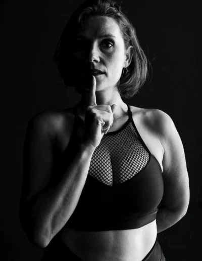 Une femme en tenue de sport pose dans un éclairage dramatique, portant un doigt à ses lèvres dans un geste de silence. Elle porte un soutien-gorge de sport en maille et regarde légèrement vers le haut, les ombres soulignant son visage et son corps sur un fond sombre.