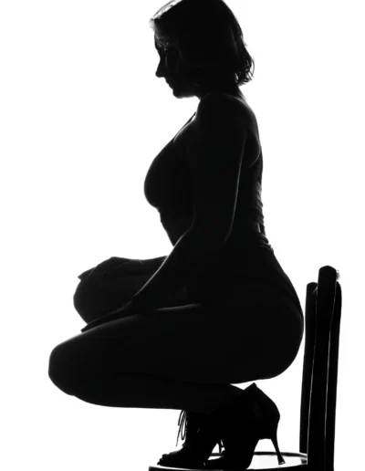 Silhouette d'une femme aux cheveux courts accroupie sur une chaise, portant un haut ajusté et des talons hauts, sur fond blanc.
