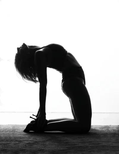 Une femme en tenue de sport prend une pose de yoga, arquant le dos et tendant les mains vers les pieds, agenouillée sur le sol, capturée dans une silhouette dramatique en noir et blanc sur un fond lumineux.