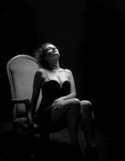 Une femme vêtue d'une robe sans bretelles est assise sur une chaise sous un éclairage dramatique et discret, regardant vers le haut avec la tête penchée en arrière, créant ainsi une atmosphère morose et artistique sur un fond sombre.