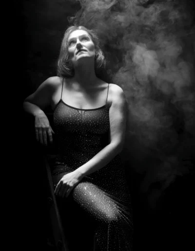 Photo en noir et blanc d'une femme vêtue d'une robe étincelante, assise sur une chaise, levant les yeux vers un faisceau de lumière entouré d'une fumée tourbillonnante dramatique dans un décor sombre.