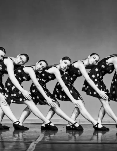 Sept danseuses vêtues de robes à motifs et de chaussures plates évoluent en ligne synchronisée, penchées en avant et les bras tendus, sur un fond de scène uni, dans une photo en noir et blanc.
