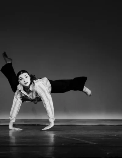 Un danseur exécute un mouvement dynamique sur scène, en équilibre sur ses mains, une jambe tendue vers le haut et l'autre pliée. La scène est en noir et blanc, avec un éclairage dramatique mettant en valeur le danseur.