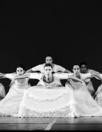 Six femmes vêtues de jupes et de robes blanches fluides posent en ligne sur scène, les bras tendus et penchés vers l'avant dans une formation de danse synchronisée, sur un fond sombre uni. L'image est en noir et blanc.