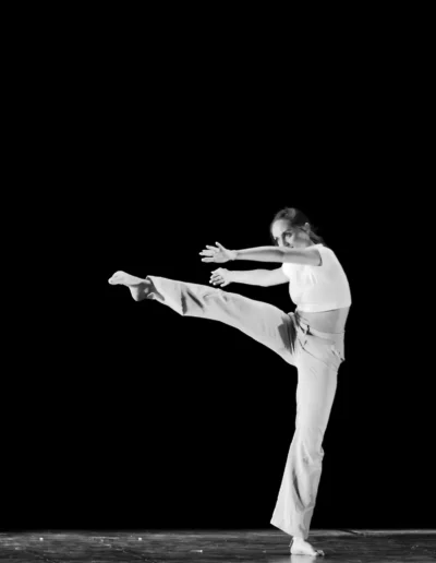 Un danseur vêtu d'un vêtement de couleur claire exécute un high kick sur scène sur fond noir, avec une jambe étendue et les bras tendus.