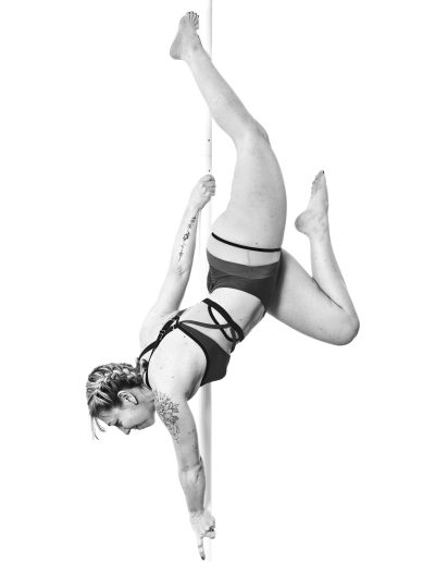 Une personne en tenue de sport effectue un mouvement acrobatique sur un poteau vertical, suspendue à l'envers, une jambe accrochée et un bras tendu vers le poteau, sur un fond blanc uni.